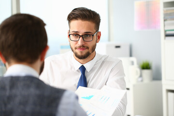 Stylish gentleman in glasses looking at male coworker and smiling while man holding financial graph