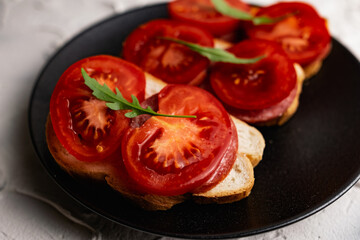 Three sandwiches with fresh and bright red tomatoes and smoked sausage, decorated with a leaf of green arugula on a matte black plate and a white stone background