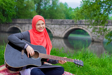 Pretty young Muslim girl trying to play acoustic guitar on the river bank