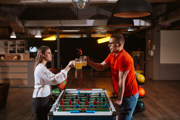 A beautiful caucasian girl and a man with glasses play table football in the playroom and drink...