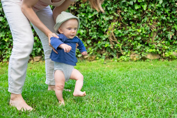 Lovely baby girl in hat and blue shirt doing first steps with help of mother. Young pretty mom holding daughter on grass. First barefoot steps, garden and sunny days concept
