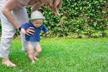 Adorable baby girl in hat and blue shirt doing first steps with help of mother. Young mom holding daughter on grass. First barefoot steps, garden and sunny days concept
