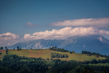 Fototapeta premium Jamnik church on hilltop with Alps mountains in background. Hill slope covered with grass and trees. Unique perspective of this famous picturesque church in Slovenia