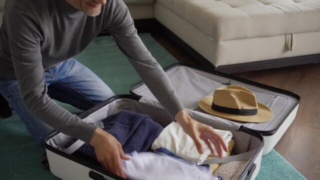 Tracking tilt up shot of senior man sitting on floor at home and packing luggage for vacations. Happy man, closing suitcase putting on straw hat on it and thinking dreamily