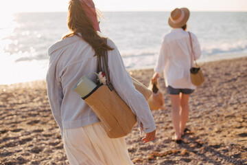 Two young beautiful women female friends going to have summer picnic on a beach at sunset.
