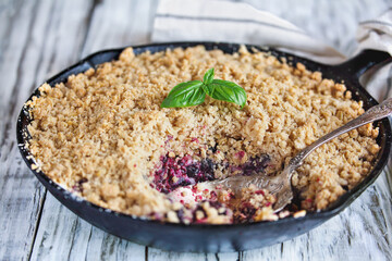 Sweet homemade Blackberry and Blueberry Basil Cobbler baked in a cast iron pan and topped with a golden oatmeal crisp over a rustic white wood table. Selective focus with blurred background.