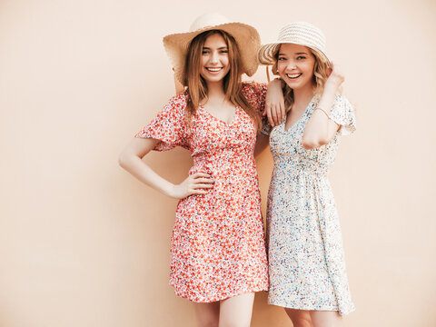 Two young beautiful smiling hipster girls in trendy summer sundress.Sexy carefree women posing on the street near wall in hats. Positive models having fun and hugging
