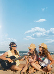 Friends on the beach a sunny summer day. They are laughing sitting on the sand and playing the guitar