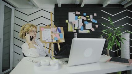Young woman talking on phone and sitting with legs on table in office