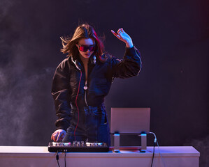 Stylish girl DJ in glasses headphones and a bomber mixes music dancing at a party in the neon light...