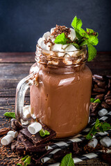 Chocolate milk shake with sauce, marshmallows and grated chocolate, decorated with mint. On old rustic wooden table with a lot of chocolate pieces, cocoa beans and chocolate shavings