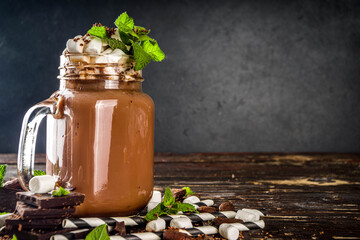 Chocolate milk shake with sauce, marshmallows and grated chocolate, decorated with mint. On old rustic wooden table with a lot of chocolate pieces, cocoa beans and chocolate shavings