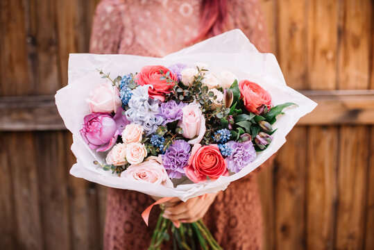 Very nice young woman holding huge beautiful blossoming bouquet of fresh carnations, roses, muskari, hyacinths, ranunculus in coral, pink, purple, blue colors on the grey background