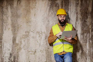 Young bearded contractor leaning on concrete wall and checking on materials on laptop.