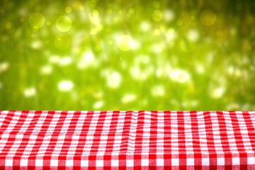 Empty table background. Empty wooden deck table covered with a red checkered tablecloth in front of abstract blurred fresh colorful green summer backdrop. Space for your food