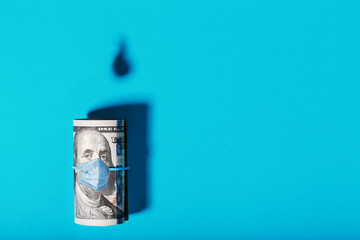 A roll of American bills with a virus mask, with the shadow of oil barrels and a drop of oil on a blue background.