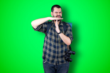 Closeup portrait serious young traveler bearded man showing loser sign on forehead looking at you with disgust at camera isolated green studio background. Negative human emotion facial expression