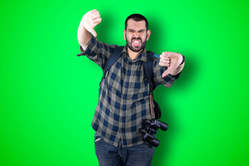 Naklejka premium Closeup portrait serious young traveler bearded man showing loser sign on forehead looking at you with disgust at camera isolated green studio background. Negative human emotion facial expression