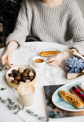 Very nice young woman sitting in a cafe, having a cup of espresso coffee coffee with some fresh eclairs, chocolate candies, eucalyptus, blue hyacinths bouquet and cappuccino cup on the white table 