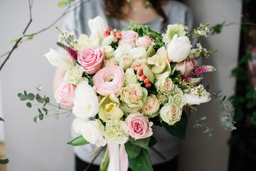 Very nice young woman holding a beautiful blossoming flower bouquet of fresh roses, ranunculus, carnations, tulips, eucalyptus in white and pink colors on the grey wall background