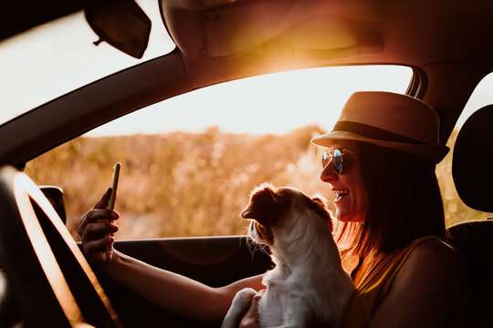 Happy young woman in a car with her dog at sunset. Taking a picture with mobile phone. Travel concept