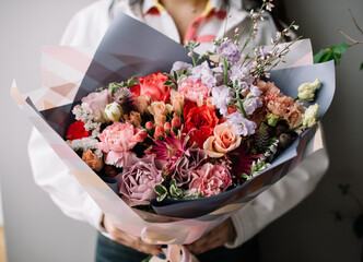 Very nice young woman holding huge beautiful blossoming bouquet of fresh roses, gerbera, carnations, eustoma, matthiola, genista, statice in red and purple colors on the grey background