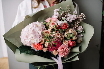 Very nice young woman holding huge beautiful blossoming bouquet of fresh hydrangea, carnations, roses, chamellacium, matthiola, ranunculus in purple and red colors on the grey background