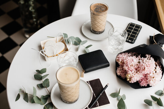 Coffee break at the cafe: two coffee cups, black leather wallet, hyacinths flower bouquet, cookie, phone and eucalyptus on the white table background