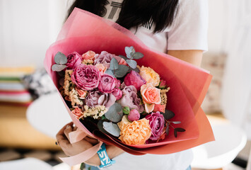 Very nice young woman holding huge beautiful blossoming bouquet of fresh roses, carnations, statice, eucalyptus in pink, purple and orange colors on the grey background
