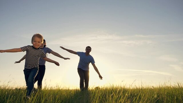 Silhouette of a happy family. Mom, dad, and son run across the field at sunset with their arms spread wide like wings and imitate the flight of an airplane.