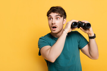 handsome shocked man holding binoculars on yellow