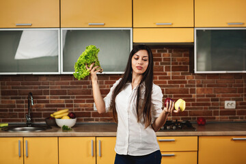 Portrait of a young woman holds two apples and lettuce in her hands. A woman standing in the kitchen at home. The concept of diet and healthy eating, cleansing the body