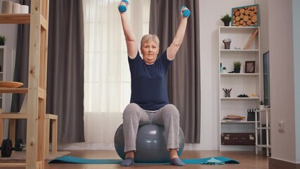 Senior woman lifting dumbbells sitting on fitness ball in living room. Old woman lifting training healthy lifestyle sport fitness workout at home with weights dumbbell activity - Powered by Adobe
