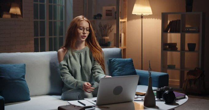 Pretty caucasian redhead girl sitting on coach looking at laptop, having a videochat or online tutorial, recording an internet vlog, cute model looking at webcamera and talking 4k footage