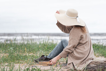 A woman dressed in a warm cardigan and a beige hat reads a book against the backdrop of the sea during a storm