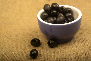 Black seedless olives in a ceramic bowl and a few scattered olives on a background of coarse-textured fabric. Close up.