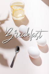 chicken eggs, teaspoon and glass of water with lemon on grey table with breakfast lettering