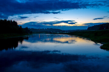 Silhouettes of trees and a bridge under a dark sunset sky that is reflected in the river.