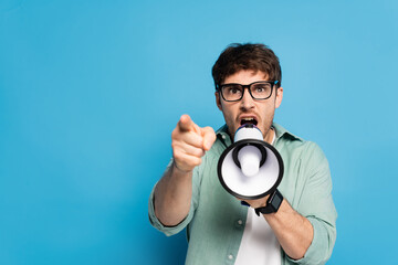 aggressive young man screaming in megaphone and pointing with finger on blue