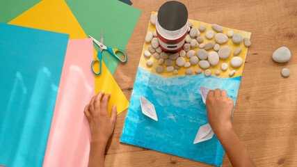 Kid hands make a lighthouse for school project at home. Top down view to wooden table with colored...