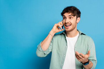 cheerful young man talking on smartphone while standing with open arm on blue