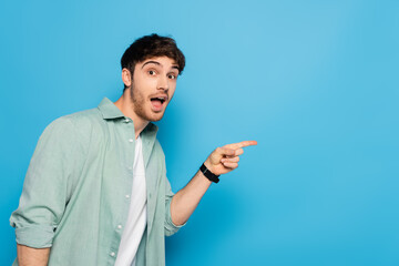 shocked young man looking at camera and pointing with finger on blue