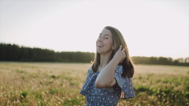 A caucasian beautiful wonan standing on the beautiful field during summer sunset and looking at the camera. The woman wears beautiful dress and. The woman is smiling. Close up. SLOW MOTION