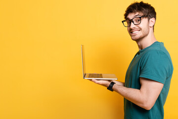 smiling young man looking at camera while holding laptop on yellow