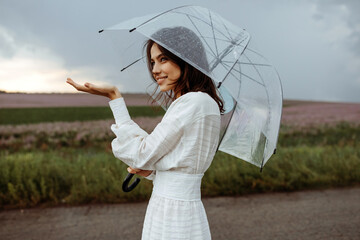 Young woman holding a clear, transparent umbrella on a stormy weather, standing in an open field,...