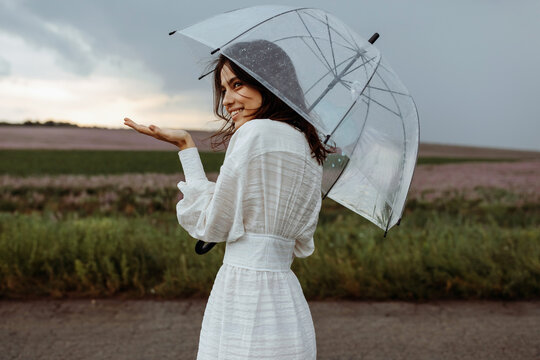 Young woman smiling, holding a clear, transparent umbrella, standing in an open field, checking the rain, on a dramatic dark blue sky background.
