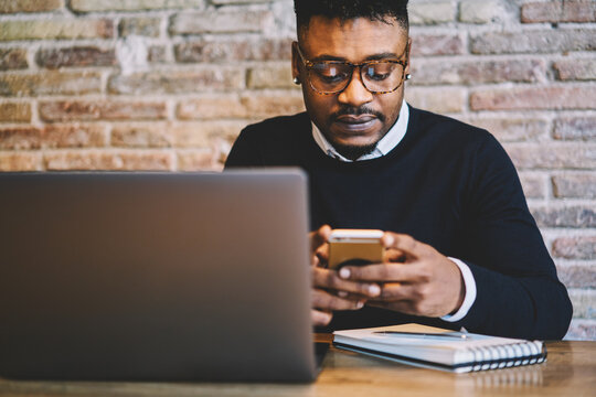 Cropped image of afro American hipster guy in eyeglasses doing shopping on internet websites on mobile phone using free internet connection. Hipster man downloading new mobile application on cellular