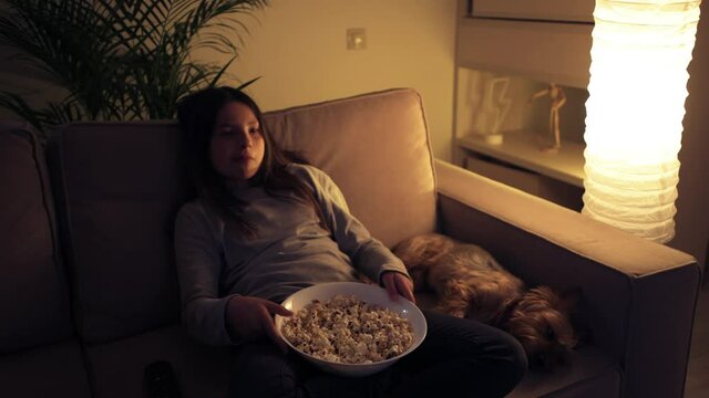 Tired girl sitting at home on the couch with her dog and watching a television.