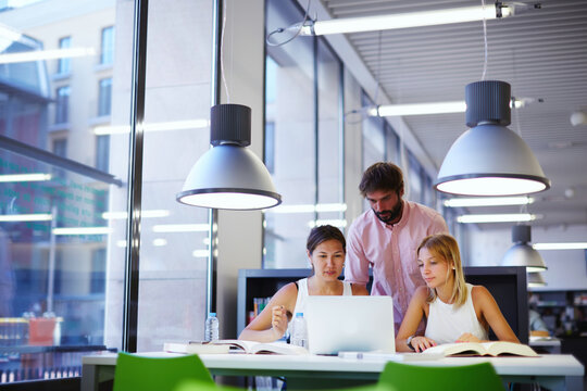 Group of international university students learning in library, three colleagues of modern work co-working space talking and smiling while sitting at the desk with laptop computer, exam preparation