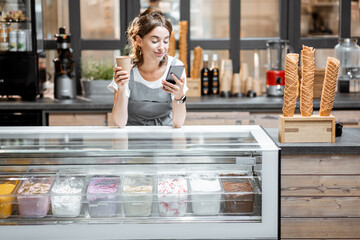 Portrait of a young and happy saleswoman with coffee and phone at the counter in ice cream shop or...